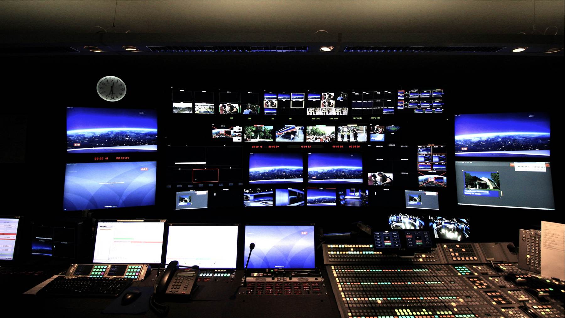 A broadcast control room with a wide wall of monitors and a mixing console